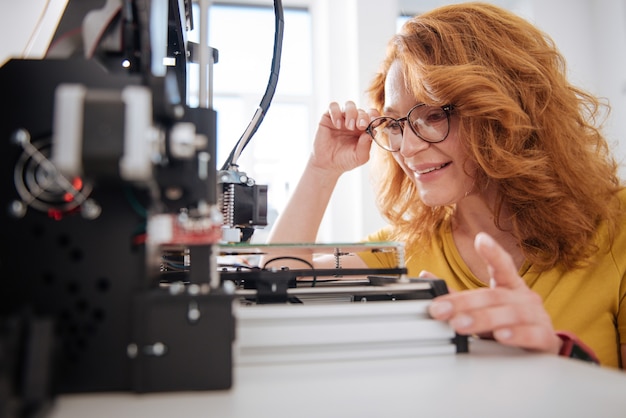 Bella donna positiva piacevole che si protende in avanti e fissa gli occhiali mentre guarda i dettagli della stampante 3d