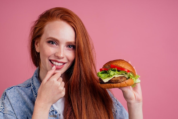 Bella donna rossa dai capelli rossi che tiene un grande cheeseburger con cotoletta di cheesebeef e lattuga al pomodoro e lecca le dita in studio sfondo rosa