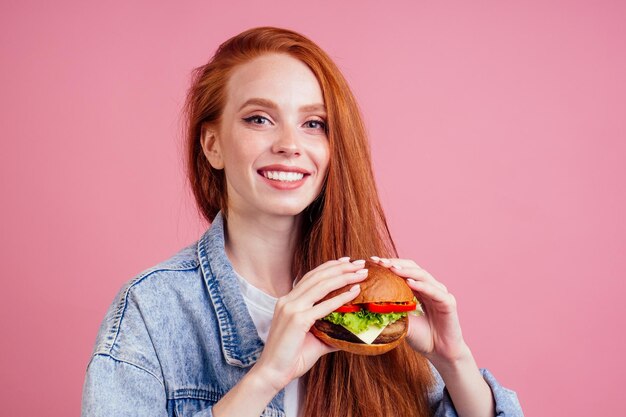 Bella donna rossa dai capelli rossi che tiene un grande cheeseburger enorme con cotoletta di manzo al formaggio e lattuga di pomodoro in studio sfondo rosa