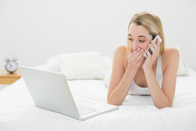 Bella donna scioccata telefonando mentre giaceva sul suo letto guardando portatile