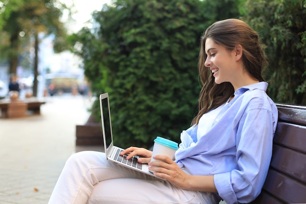 Bella donna seduta sulla panchina che tiene il caffè mentre si utilizza il computer portatile all'aperto.