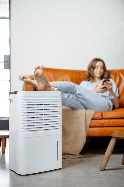 Bella donna seduta vicino al purificatore d'aria e all'apparecchio idratante vicino al divano per monitorare la qualità dell'aria nel telefono. Microclima di salute a casa concetto.