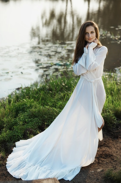 Bella donna snella in un lungo abito bianco cammina la mattina vicino al lago.