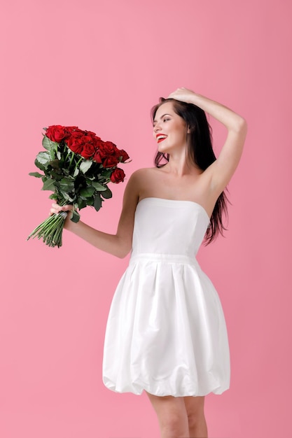 Bella donna sorpresa in un abito bianco con un grande mazzo di rose rosse su sfondo rosa. San Valentino