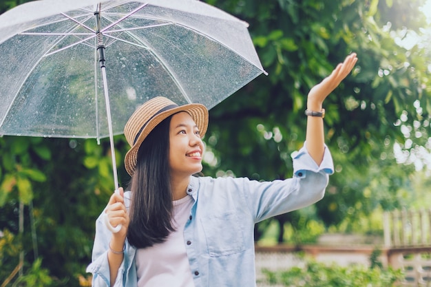 Bella donna sorridente asiatica che copre gli ombrelli sotto la pioggia con la mano che gioca goccia di pioggia nel parco