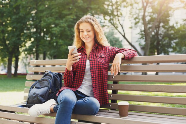 Bella donna sorridente che utilizza smartphone sulla panchina all'aperto. Tecnologia, social media, istruzione e concetto di freelance, spazio di copia