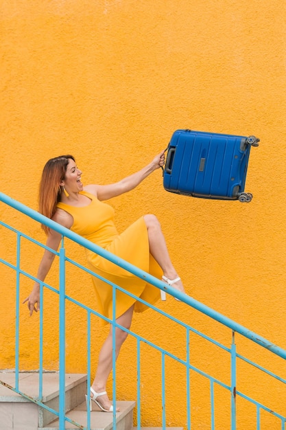 Bella donna sorridente dai capelli rossi scende con gioia le scale e agitando una valigia blu su sfondo giallo