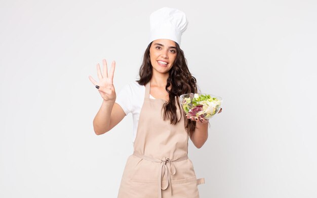 Bella donna sorridente e dall'aspetto amichevole, mostrando il numero quattro con indosso un grembiule e con in mano un'insalata