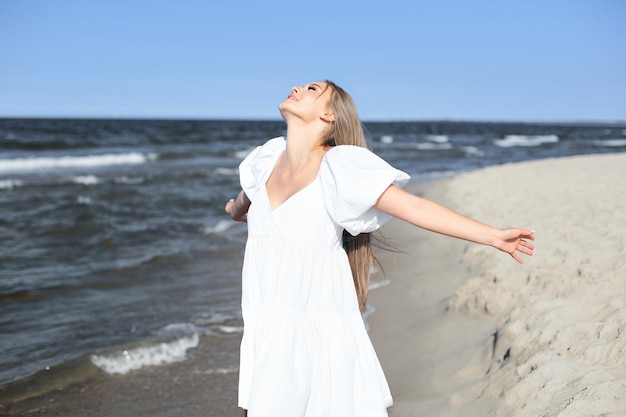 Bella donna sorridente felice è sulla spiaggia dell'oceano in un abito estivo bianco, a braccia aperte