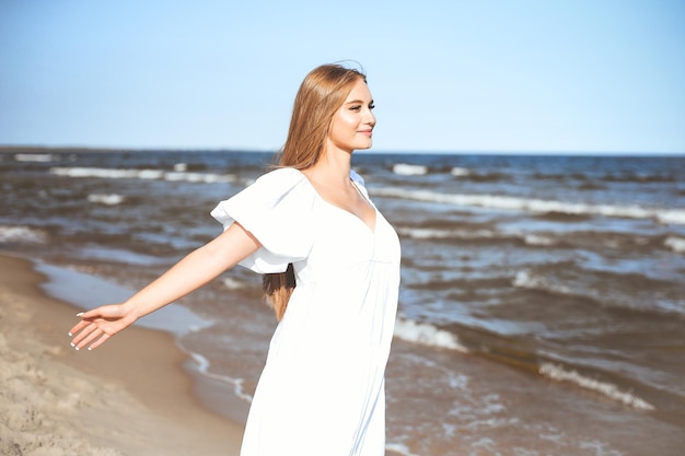 Bella donna sorridente felice è sulla spiaggia dell'oceano in un abito estivo bianco, a braccia aperte.