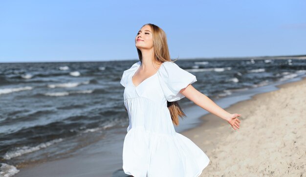 Bella donna sorridente felice è sulla spiaggia dell'oceano in un abito estivo bianco, a braccia aperte.