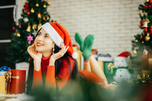 Bella donna sorridente felice in abito rosso e con indosso un cappello da Babbo Natale è sdraiata a terra circondata da scatole regalo colorate con un albero di Natale sullo sfondo che tiene in mano un tablet.