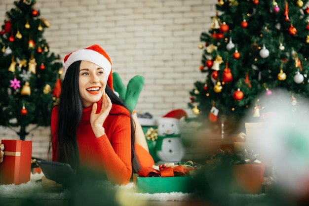 Bella donna sorridente felice in abito rosso e con indosso un cappello da Babbo Natale è sdraiata a terra circondata da scatole regalo colorate con un albero di Natale sullo sfondo che tiene in mano un tablet.