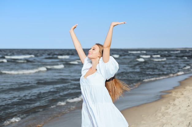 Bella donna sorridente felice sulla spiaggia dell'oceano in piedi in un abito estivo bianco, alzando le mani.