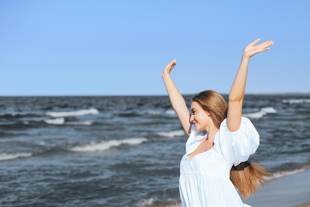 Bella donna sorridente felice sulla spiaggia dell'oceano in piedi in un abito estivo bianco, alzando le mani.