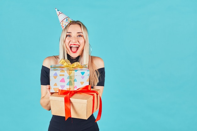 Bella donna sorridente in vestito nero e cappello di compleanno che tiene il contenitore di regalo sulla parete blu. Ragazza giovane felice sveglia che celebra la festa di compleanno.