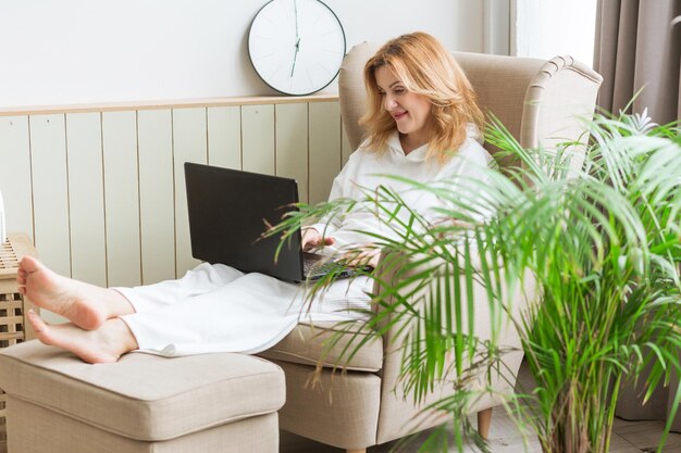 Bella donna sorridente matura che lavora al computer portatile e che si siede su una sedia grande e comoda a casa