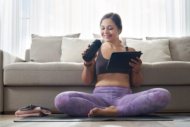 Bella donna sportiva atletica che si siede sulla stuoia di yoga con la compressa dopo alcuni esercizi e bevande