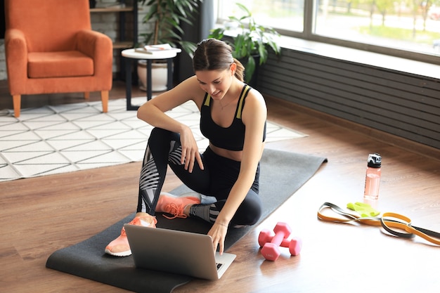 Bella donna sportiva snella in abbigliamento sportivo è seduta sul pavimento con manubri e bottiglia d'acqua e sta usando un computer portatile a casa nel soggiorno. Uno stile di vita sano. Restare a casa attività.