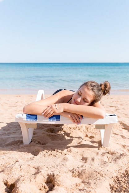 Bella donna sta prendendo il sole sul lettino in spiaggia e ha una faccia sorridente fatta di crema solare sulla sua mano.