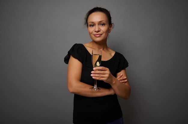 Bella donna vestita di nero, sorride guardando la fotocamera e posa su sfondo grigio muro con un flauto di champagne in mano. Concetto di celebrazione del Black Friday con copia spazio per la pubblicità