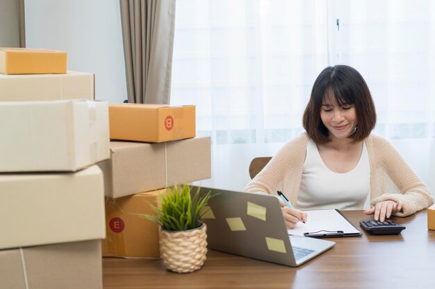 Bella e affascinante giovane donna asiatica piccola imprenditrice online che controlla i suoi ordini sul laptop mentre prepara le scatole di spedizione