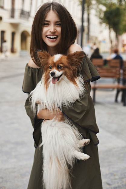 Bella e allegra ragazza modella bruna con sorriso splendente, in abito corto con un piccolo cane carino papillon sulle sue mani in posa all'aperto nel centro storico della città