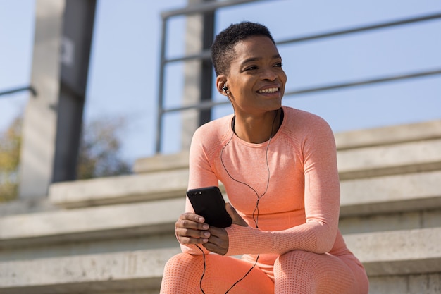 Bella e atleta di successo femminile sorridente ascolta l'audiolibro dal telefono cellulare durante il fitness mattutino e il jogging vicino allo stadio, donna afroamericana con il telefono cellulare