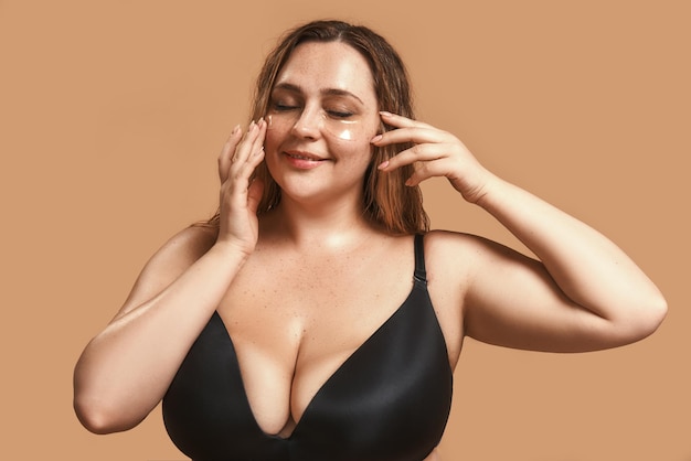 Bella e felice donna plus size in biancheria intima nera che applica cerotti sotto gli occhi e sorride mentre si trova in studio su sfondo marrone. Ritratto in studio. Donna sovrappeso