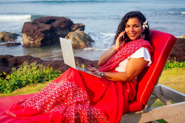 Bella e giovane donna d'affari indiana nel tradizionale sari indiano che lavora con il computer portatile dal freelancer freelance del lavoro a distanza della studentessa seaasia seduto sulla spiaggia utilizzando un telefono smartphone