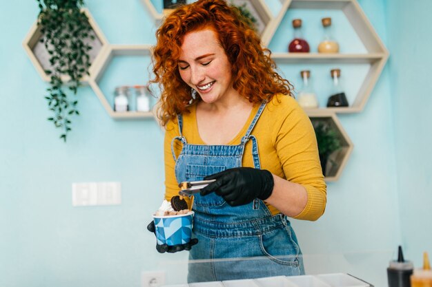 Bella e positiva donna rossa allo zenzero che sorride e lavora in una gelateria artigianale. Lei prepara e serve deliziosi dolci.