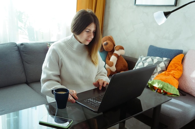 Bella e seria giovane donna seduta sul divano con peluche e lavorando con un laptop a casa con una tazza di caffè il concetto di lavoro a distanza a casa