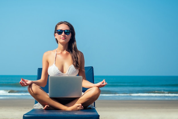 Bella e sexy giovane donna seduta nella posa del loto e meditando su un lettino con un laptop in riva al mare