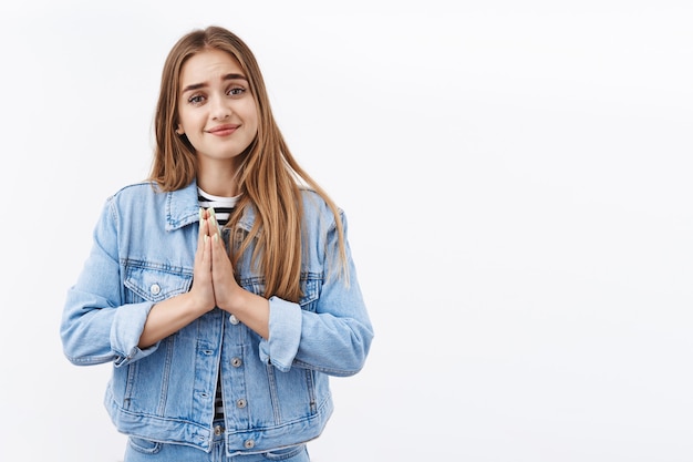 Bella e speranzosa ragazza bionda gentile che implora favore, sorride graziosamente e stringe le mani insieme in preghiera, supplica, dicendo per favore con un viso sincero e carino, ha bisogno di aiuto, in piedi sul muro bianco