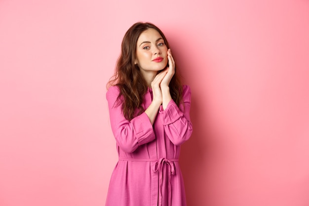 Bella e tenera donna che tocca il suo viso idratato perfetto con trucco luminoso, tenendo la mano sulla guancia, guardando la telecamera, in piedi sopra il muro rosa.