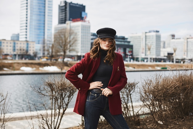 Bella ed elegante giovane donna in berretto, cappotto rosso e occhiali da sole in posa davanti al muro beige.