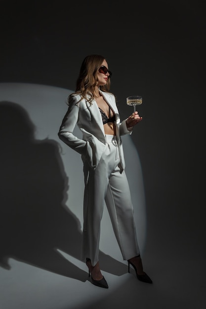 Bella elegante giovane donna chic con occhiali da sole in un abito alla moda bianco con top blazer e tacchi con un bicchiere di champagne su uno sfondo scuro in studio Lady celebra un evento