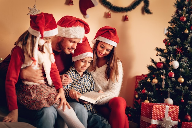 Bella famiglia sorridente di quattro persone con cappello da Babbo Natale che legge un libro a casa vicino a un albero di Natale.