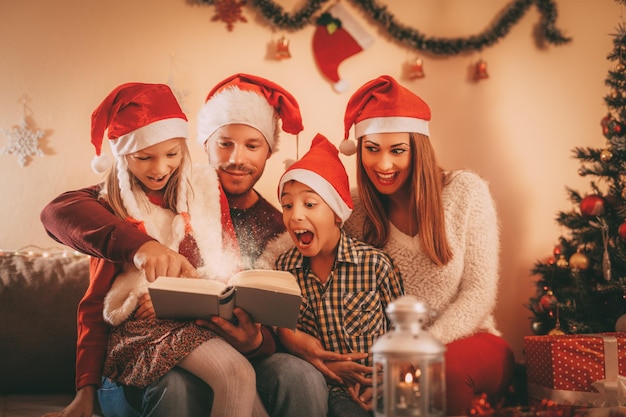 Bella famiglia sorridente di quattro persone con il libro di lettura del cappello di Babbo Natale a casa.