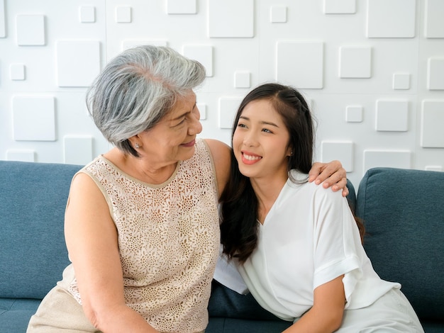 Bella felice donna asiatica figlia sorridente mentre guarda la sua anziana madre capelli bianchi con abbracciare insieme mentre si siede sul divano su sfondo bianco nel soggiorno a casa Concetto di famiglia felice