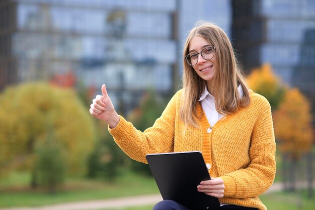 Bella felice giovane studentessa universitaria o studentessa positiva con gli occhiali è seduta nel parco, campus universitario con il suo tablet, studiando online con gadget, mostrando il pollice in su, come un gesto