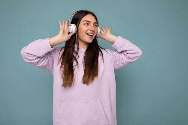 Bella felice sorridente giovane bruna persona di sesso femminile che indossa una felpa con cappuccio viola chiaro isolata su una parete di sfondo blu che indossa cuffie bluetooth bianche ascoltando musica fresca e guardando al lato