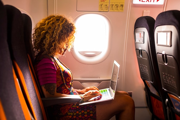 bella femmina con i capelli ricci seduta all'interno dell'aereo pronta al volo per la prossima destinazione usa un laptop durante il viaggio