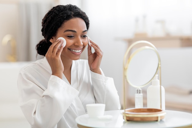 Bella femmina nera guardando lo specchio e la pulizia della pelle con tamponi di cotone