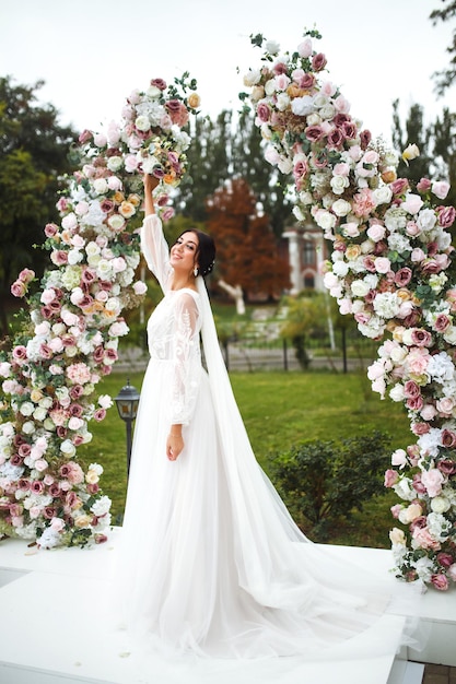 Bella fidanzata in abito da sposa bianco con bouquet vicino all'arco nuziale di fiori