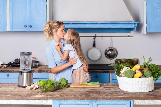 Bella figlia che bacia la giovane madre mentre si cucina insieme in cucina, preparando insalata per colazione, cesto di verdure fresche e frullatore sul tavolo, cibo vegetariano, alimentazione sana