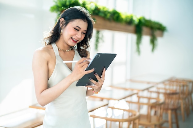 Bella freelance asiatica modello di moda donna d'affari di successo persone in abito bianco senza maniche estivo utilizzando il lavoro casual con tablet per la navigazione in internet chat e blog in caffetteria