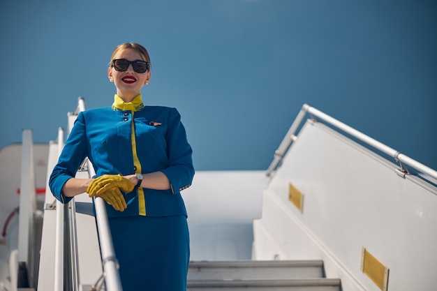 Bella giovane assistente di volo in occhiali da sole che guarda la macchina fotografica e sorride mentre mette la mano sul corrimano delle scale dell'aereo