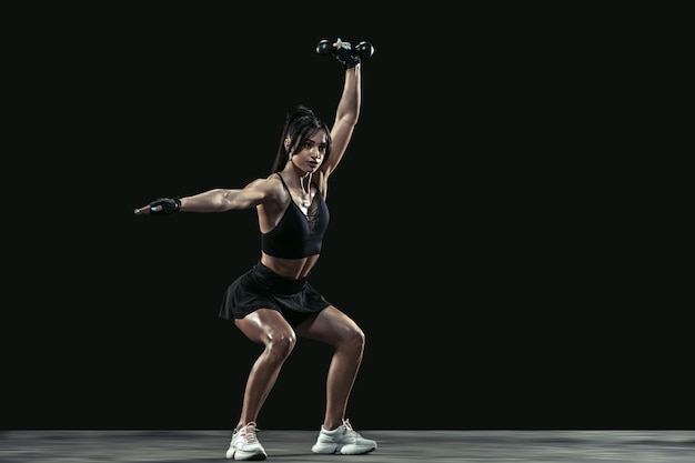 Bella giovane atleta femminile che pratica su sfondo nero studio, ritratto a figura intera. Modello bruna dalla vestibilità sportiva con pesi. Body building, stile di vita sano, concetto di bellezza e azione.