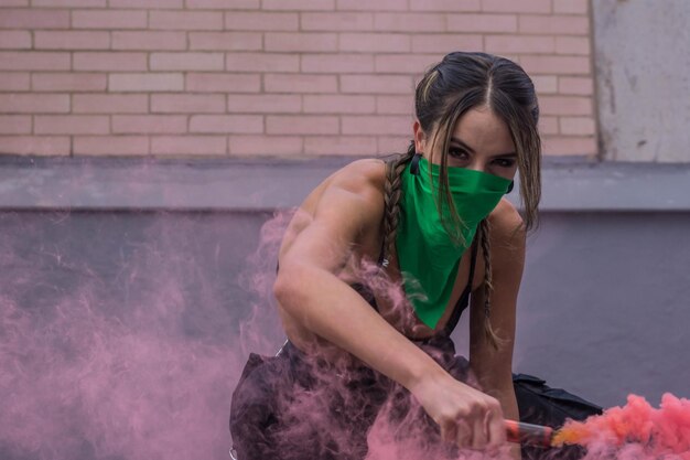 Bella giovane attivista femminista vestita di nero con la faccia coperta da una bandana verde e con in mano fumogeni rossi luminosi. Concetto femminista